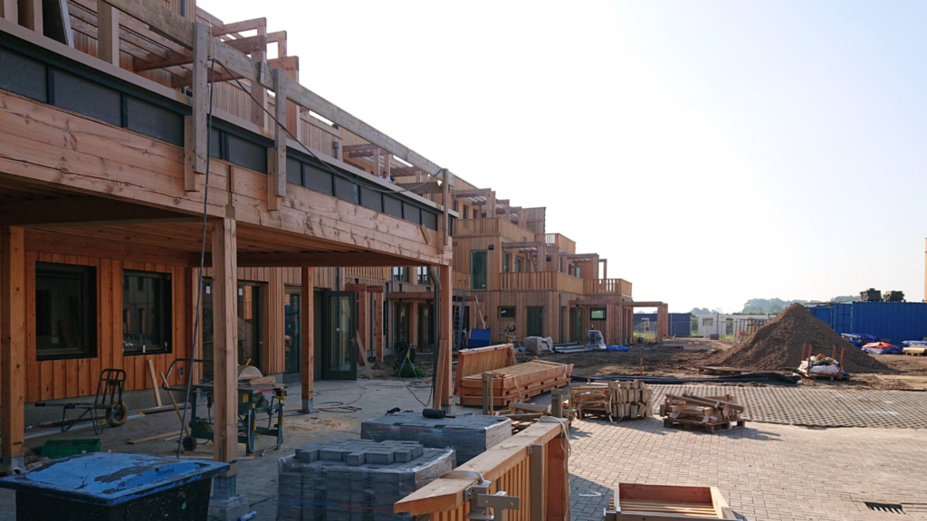Een rij woningen in aanbouw, waarbij een groot deel van de houten constructie al staat. Op de voorgrond bouwmaterialen zoals houten onderdelen van de woningen, straatstenen en werkgereedschap zoals een zaagmachine. Verderop ligt nog een bult straatzand.