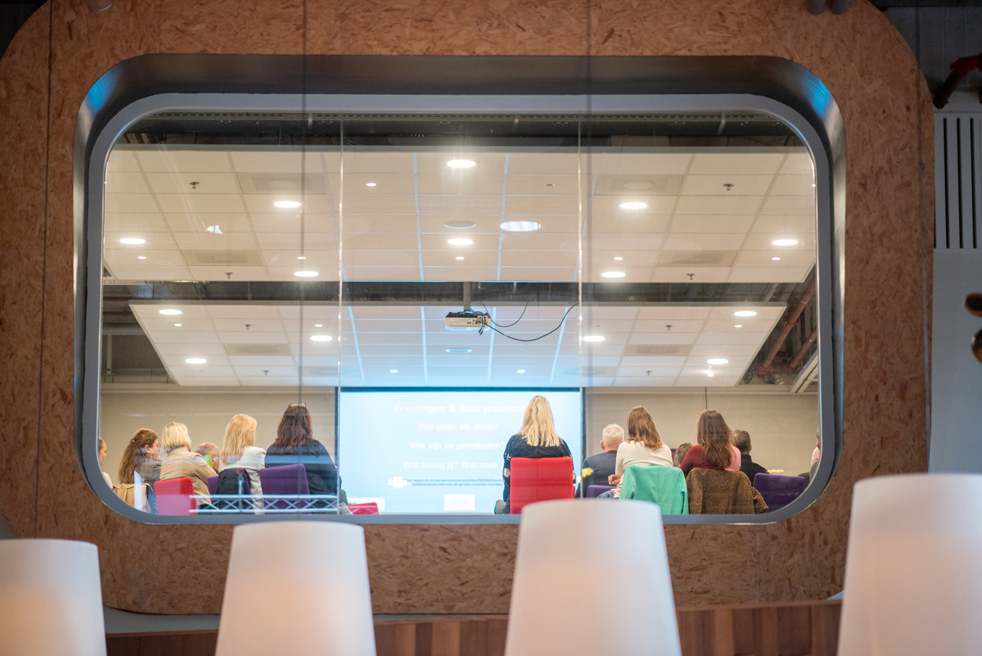 Door een raampje zie je mensen zittend in een bijeenkomst, kijkend naar een scherm