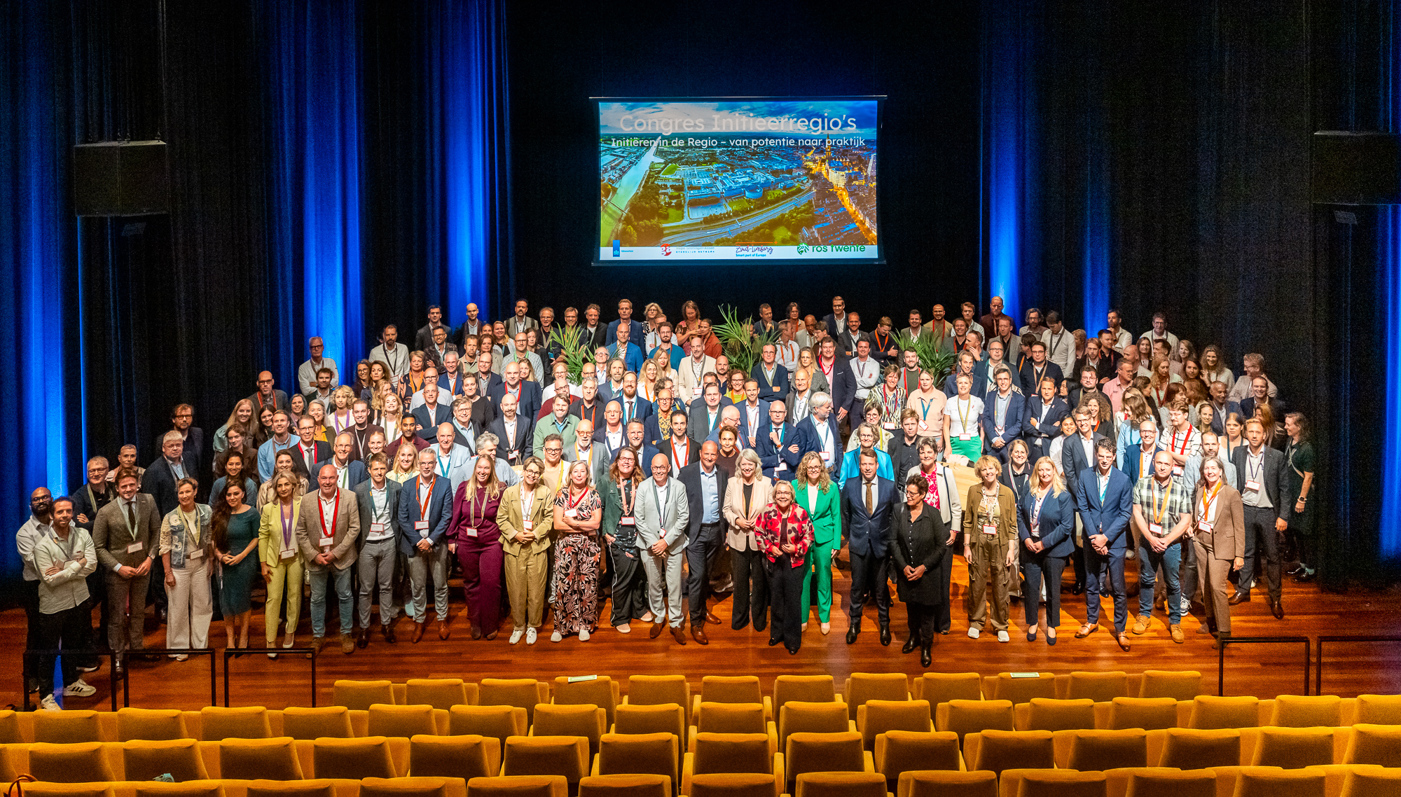 Groepsfoto met tientallen deelnemers van de bijeenkomst uit alle regio's samen op het podium. Achter hen een presentatiescherm en vooraan in beeld de lege stoelen waar de deelnemers eerst zaten.